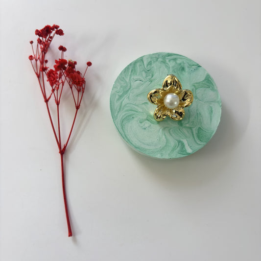 Green marbled circular object with gold flower and pearl center next to red dried flowers on a white background