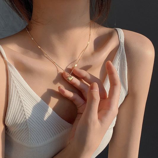 Woman wearing a delicate gold necklace with a heart pendant, set against a neutral background.