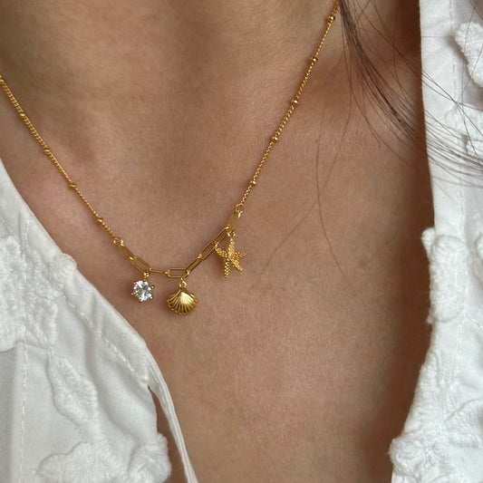 Gold necklace with starfish, shell, and crystal pendant on a person wearing a white top.