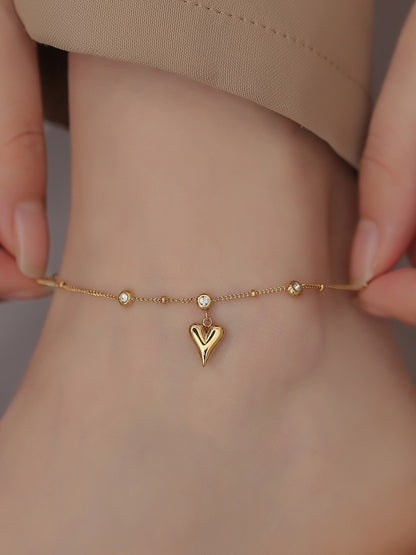 Gold anklet with heart charm on a wrist against a neutral background