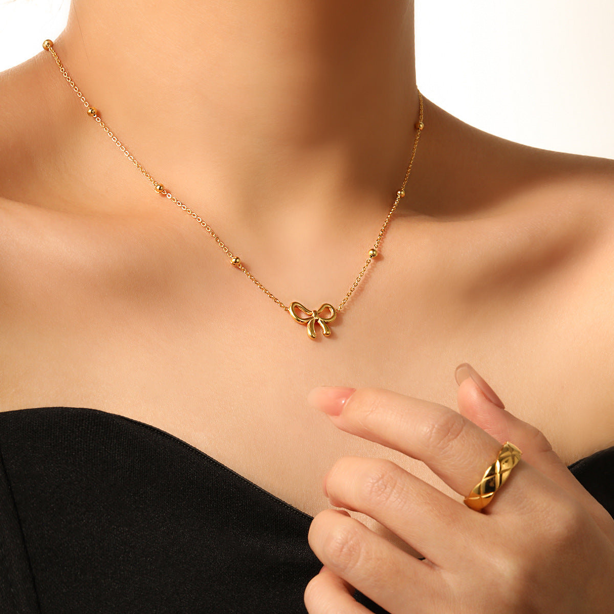 Gold necklace with a bow pendant worn by a person, with a hand wearing a matching gold ring.