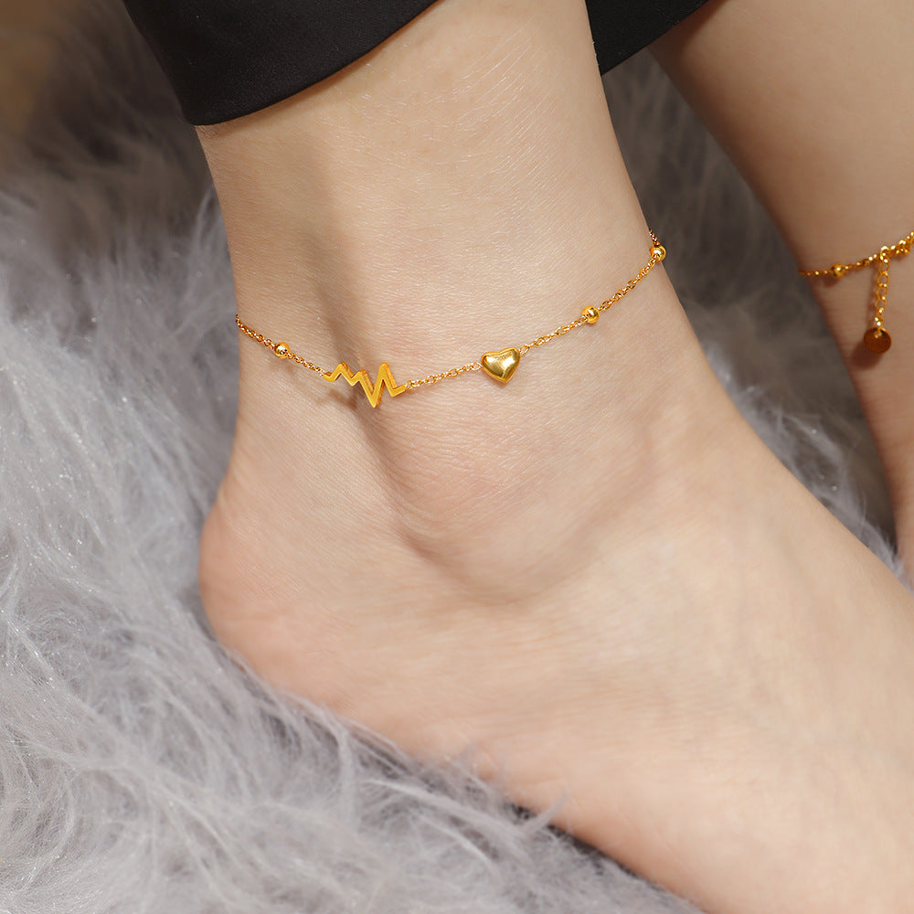 Gold anklet with heart and wave design on a person's ankle against a gray fur background