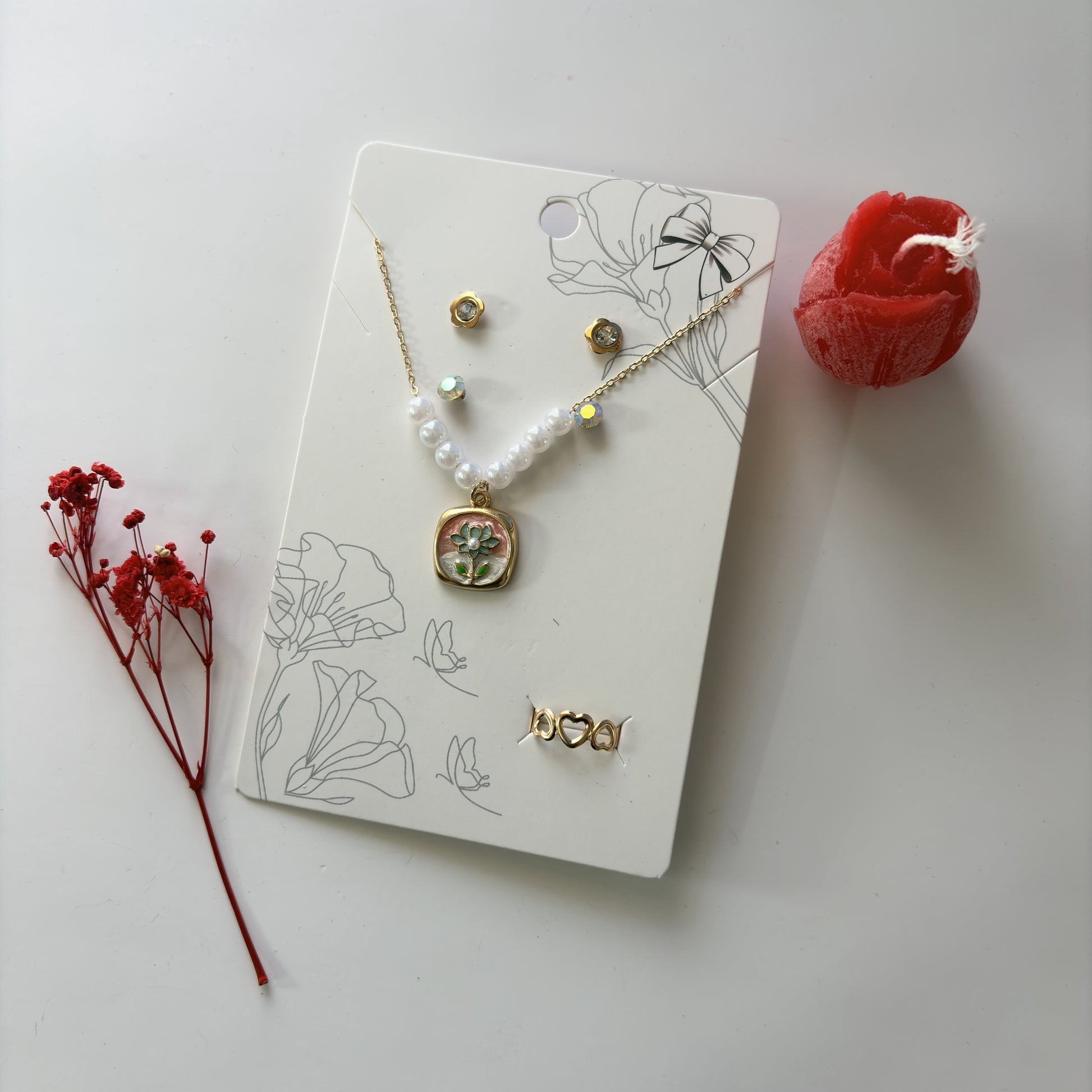 Jewelry set with necklace, earrings, and pendant on a decorative card with floral designs, accompanied by red flowers.