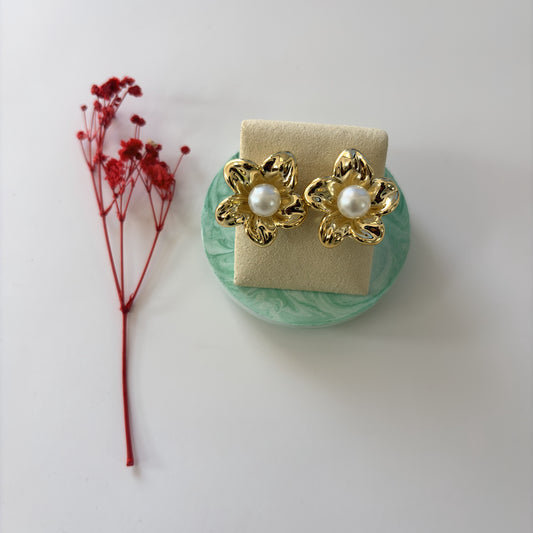 Gold floral earrings with pearl centers on a green stand, accompanied by red flowers on a white background.