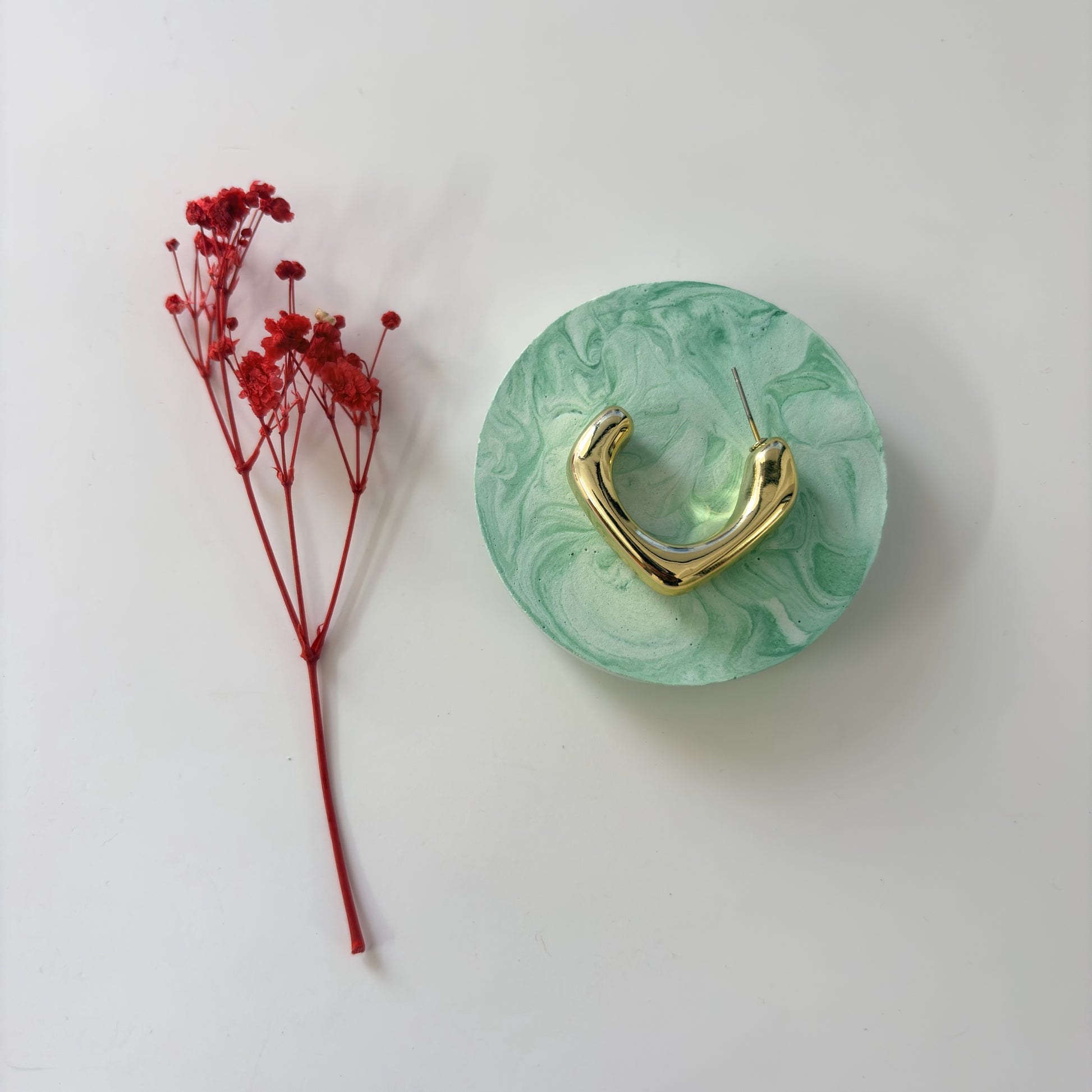 Gold hoop earring on a green marbled disc with red dried flowers on a white background
