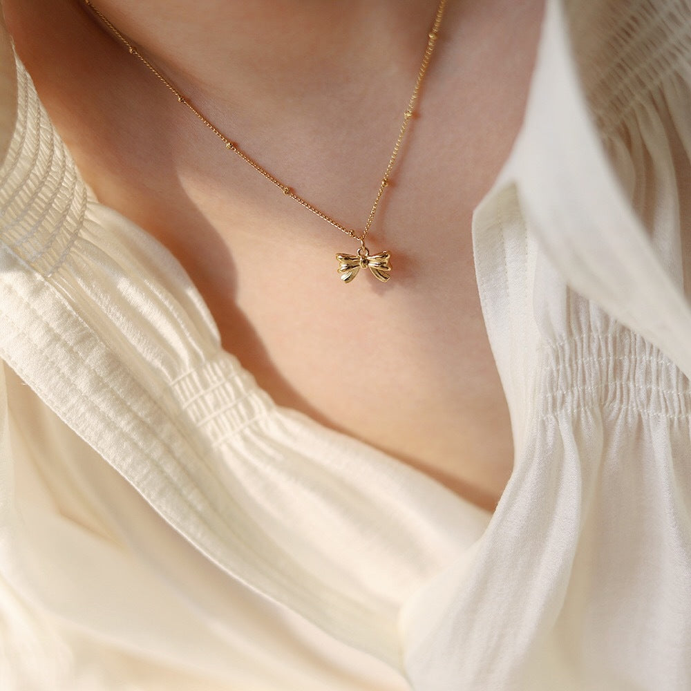 Gold necklace with a bow pendant worn by a person in a white blouse.