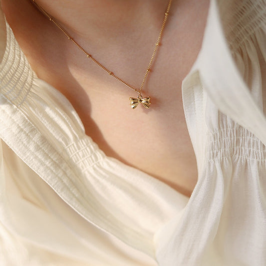 Gold necklace with a bow pendant worn by a person in a white blouse.